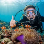 Happy beginner diver with PyraSafari instructor giving thumbs up underwater, surrounded by clownfish in Sharm El Sheikh.
